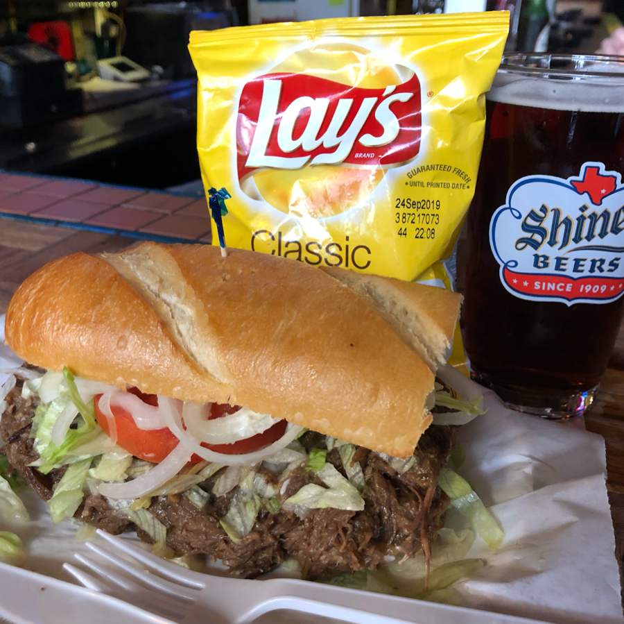 A po-boy sandwich on a paper plate with a bag of chips and a glass of beer
